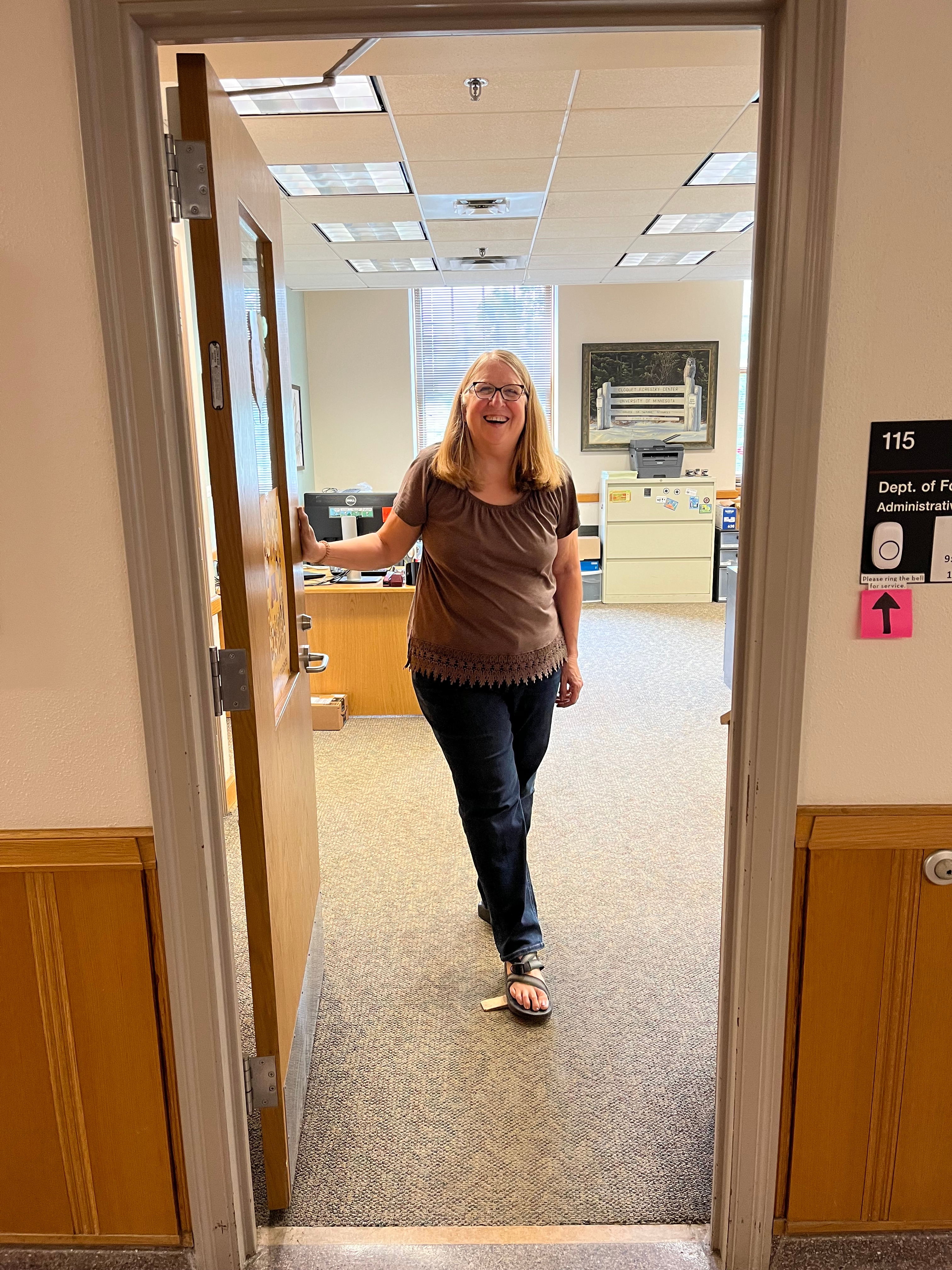 A woman with straight, shoulder-length blond hair and black-rimmed glasses stands smiling in an office doorway as she opens the door. She's wearing jeans and a brown short-sleeved top with brown lace decorating the hem.