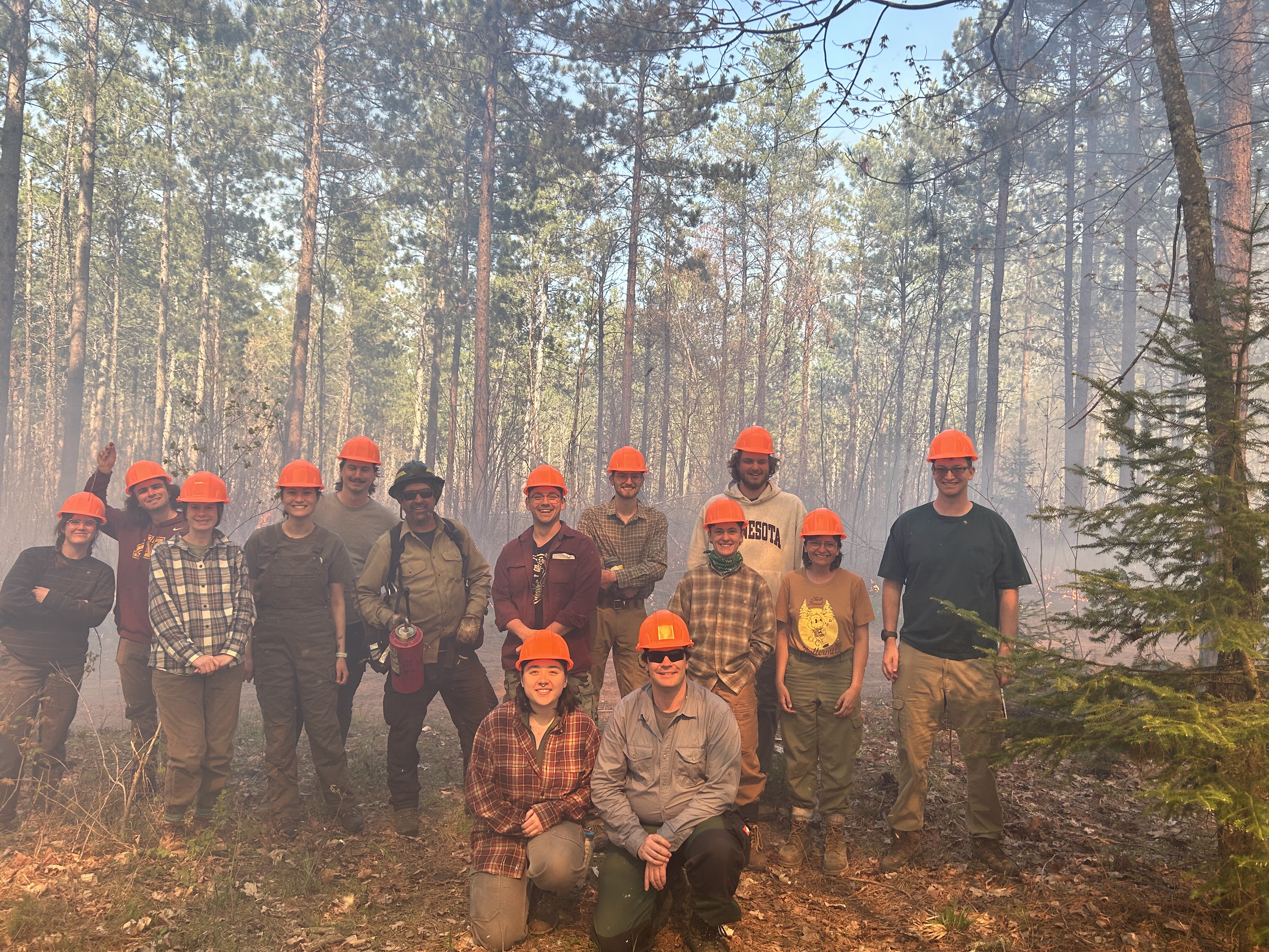 A group of young people in field gear and orange hard hats smile in a forested area near where prescribed burns are happening. An amber glow is cast across their faces. In the center of the group is a man with a drip torch, who is a member of the professional prescribed burn team at the CFC.