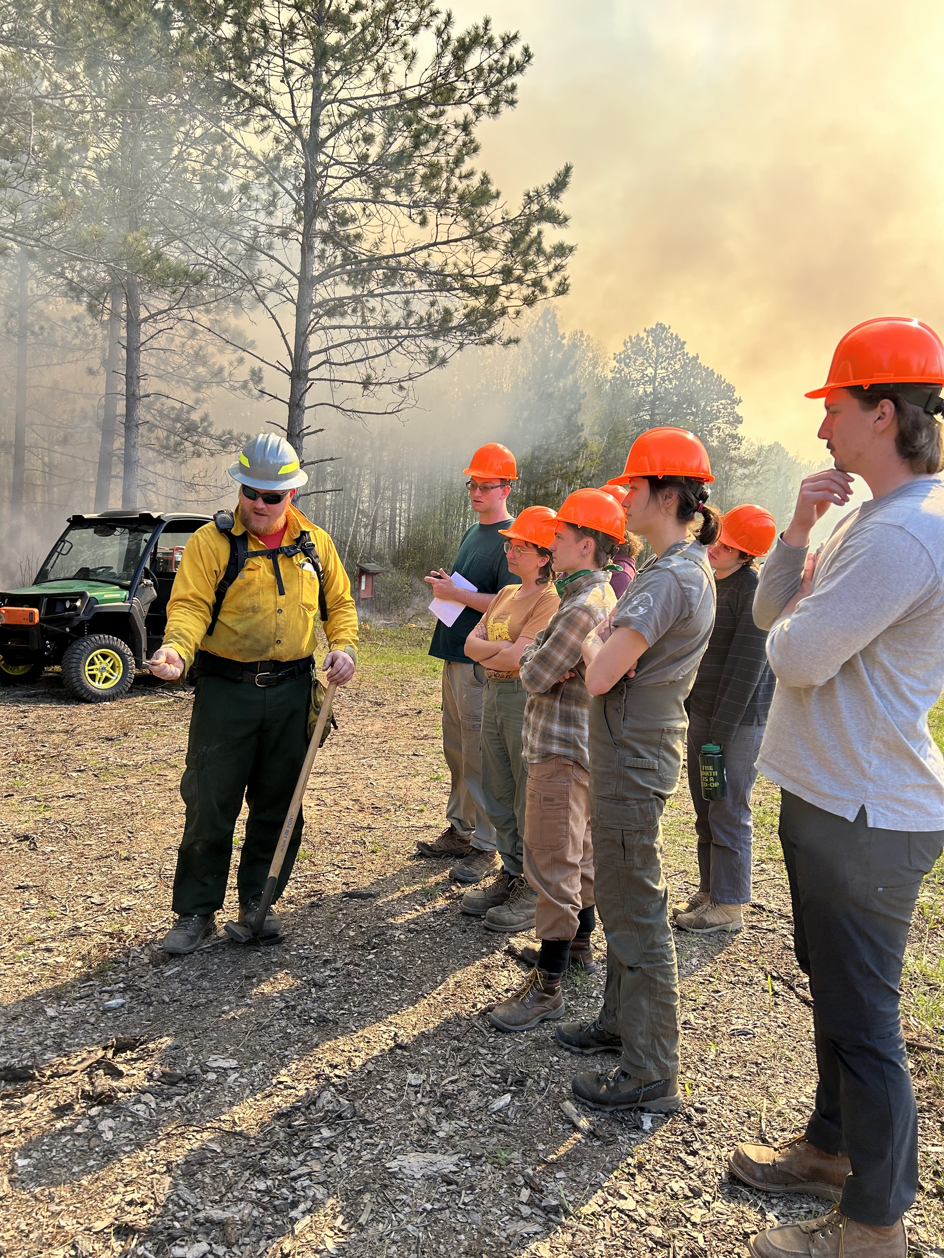 A member of the prescribed burn team, holding equipment, explains what's happening at the burn to UMN students. Everyone wears field gear and hard hats. The sky behind them is smoky with a golden hue.