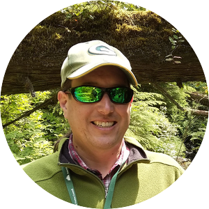 profile photo of Mike Dockry, a Native American man in a green fleece with a hat and sunglasses standing in front of a green forest