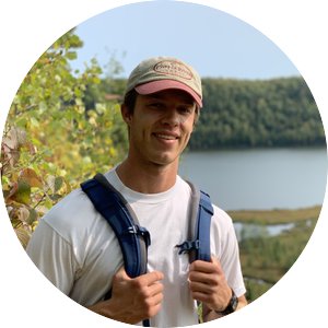 A young Caucasian man in a baseball cap, white T-shirt, and backpack, smiles in front of a lake.