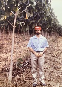 Bolstad stands next to a row of young trees. He's much younger, with a brown beard, red baseball cap, a short-sleeved button-down shirt, khaki pants, and tennis shoes.