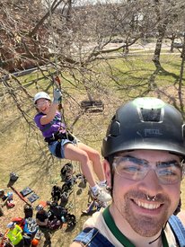 Theoren Keenan, TAG member, takes a selfie while climbing up the cottonwood tree. Theoren's face is closest to the camera and another student dangles by ropes in the background. Both are smiling and are wearing helmets and harnesses.