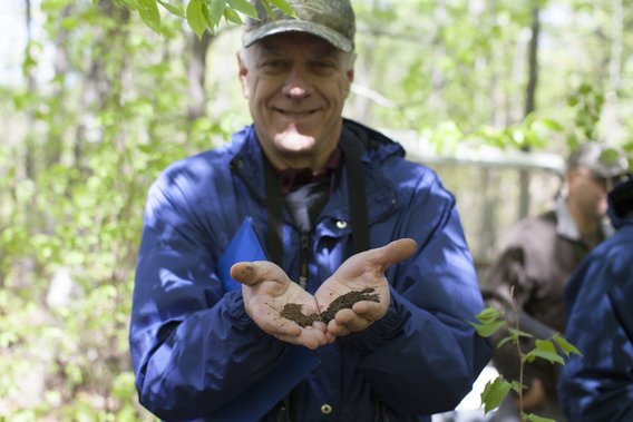 Charlie Blinn, wearing a tan baseball cap and blue jacket, holds out his hands, which are cupping some dirt. He is an older Caucasian man with white hair and a big smile. 