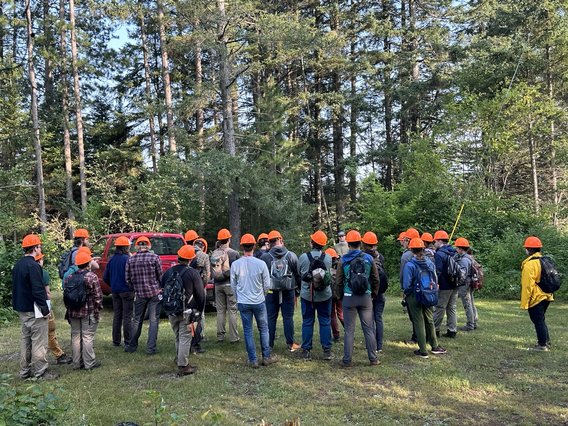 A group of about 24 students have their backs turned to the camera as they listen to instructions. They are in a clearing facing a wooded area. All wear orange hardhats and long-sleeved shirts and pants. A red truck is in the background.