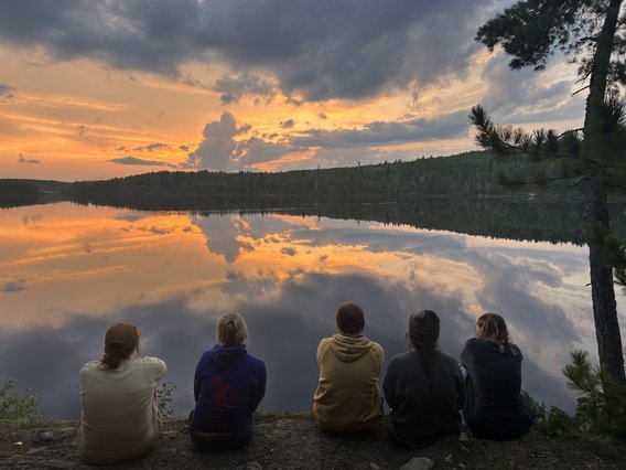 A group of five people are seated on the edge of a lake watching a sunset. They are dressed in sweatshirts. The sky reflects in the water below and bursts with a splash of orange and pink light through some gray clouds.