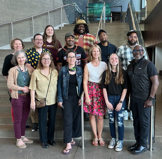 A group of 13 people smile together on a staircase. They are different ages, ethnicities, and genders.