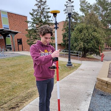 Cole Kiernan, a white man with curly brown hair and a maroon sweatshirt holds a red and white instrument pole