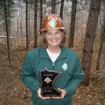 Jan Bernu, a white woman in a green coat holds an award in front of a forest