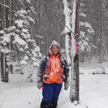 Jan Bernu, a white woman in blue coat and pants with an orange vest stands in a snowy forest