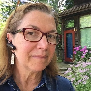 image of Kristen C. Nelson, a white woman with blond hair and red glasses wearing a bluetooth earpiece