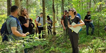 Catherine Glenn-Stone, a white woman in a black shirt and pants smiles at classmates in a green forest