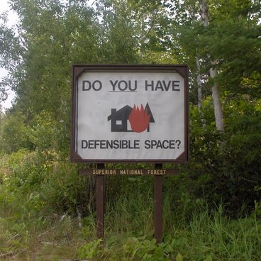 image of a white sign in front of the superior national forest that says 'Do you have defensible space?' with a graphic of a burning house