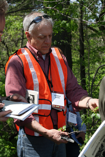 Charlie Blinn wears an orange safety vest, a plaid shirt, and jeans in the woods. He studies a low tree branch while another person takes notes beside him.