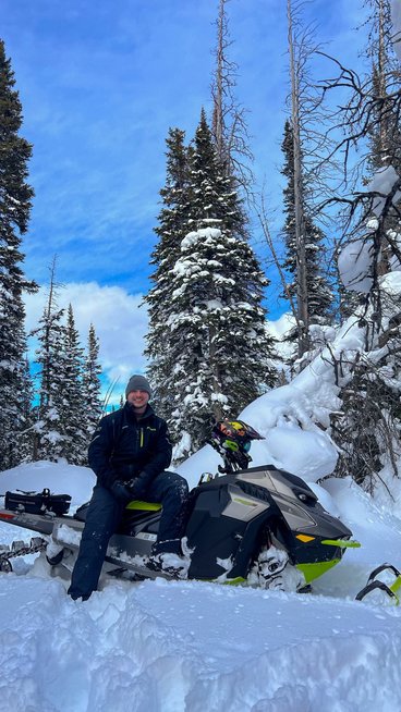 Adam Langer, a young Caucasian man, rests on a snowmobile mid-trail. He's wearing dark snowmobile gear and is in front of tall, snow-covered evergreens and a bright blue sky. 