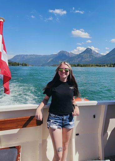 Kylee Gregory, a young Caucasian woman, smiles brightly while leaning against the side of a boat on the water. Behind her is deep blue water, a bright blue sky with a few white clouds, and mountains. Gregory has long straight hair, big sunglasses, and is wearing a T-shirt and jean shorts.
