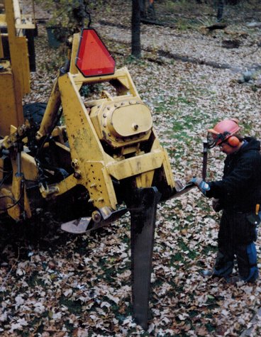 Steve Cook stands to the right of a yellow vibratory plow cutting roots between oak trees. He's is wearing an orange hardhat and ear muffs and is looking down while the machine works.