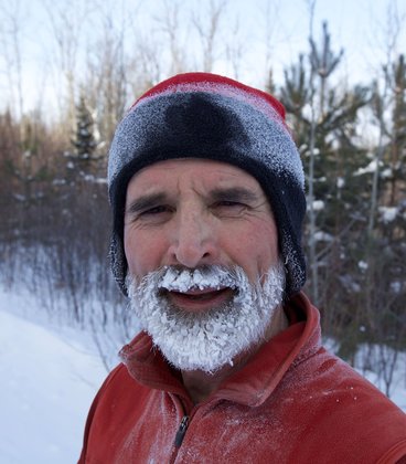 Dr. Paul Bolstad smiles at the camera in winter. He's a Caucasian man whose full beard is covered in white frost. Bolstad wears a red winter coat and a red-and-blue hat. Behind him are snow-covered trees.