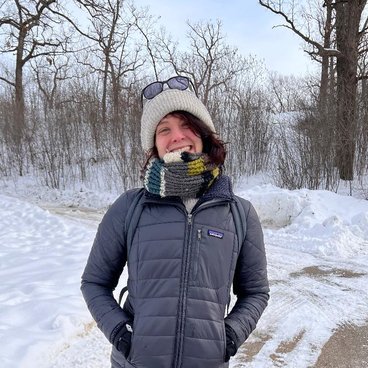 A Caucasian woman smiles while bundled in a coat, hat, and scarf in winter. She has sunglasses resting on top of her hat. Behind her is snow, bare trees, and a clear sky.