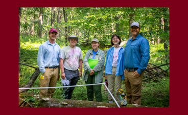 In a photo taken in the woods, five people dressed in field gear stand smiling. One person is wearing a UMN cap.