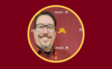 Mike Dockry smiles in front of a UMN CFANS banner in maroon and gold. He wears a maroon shirt, black glasses, and a curled moustache.