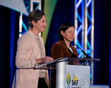 Marcella Windmuller-Campione wears a pink blazer and smiles as she speaks at a lectern on a stage. She has short brown-and-gray hair. At right is another woman with chin-length brown hair and wearing a brown corduroy blazer. The lectern has the 2023 SAF National Convention logo on it.