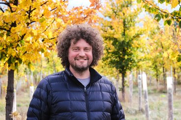 A light-skinned man with a big poof of curly brown hair and a short beard smiles in an elm nursery. It is autumn and the trees are various shades of fall colors. The man wears a thin black puffer coat. 