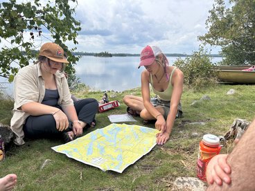 Two young women study a map while seated on some grass besides a lake. The woman on the left is wearing a brown baseball cap, a short-sleeved shirt, and long black pants. The woman on the right is wearing a red-and-white mesh baseball cap, a tank top, and shorts. There is a box of snacks in the background and another person's arm shows in the bottom right corner of the photo. There is the bow of a canoe in the background.