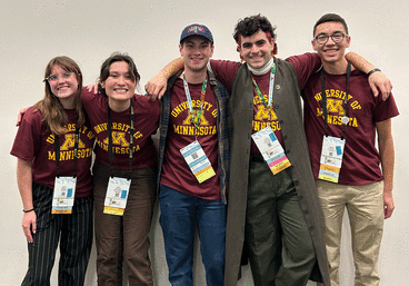 Five undergraduates wearing maroon-and-gold UMN tees and convention badges stand in front of a white wall with their arms around each other and smiling. They are of different genders and ethnicities.