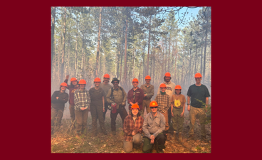 A group of young people in field gear and orange hard hats smile in a forested area near where prescribed burns are happening. An amber glow is cast across their faces. In the center of the group is a man with a drip torch, who is a member of the professional prescribed burn team at the CFC.