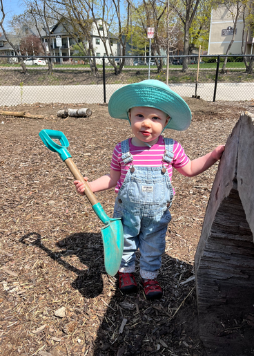 A toddler in overalls and a sun hat holds a shovel that's just her size.