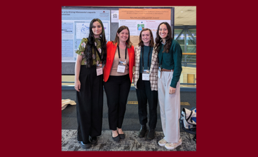 Four women stand in front of a science poster in a conference hall.