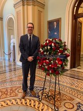 Photo of Jake Donnay, a white man with glasses wearing a suit standing next an arrangement of flowers in the U.S. Capital Building